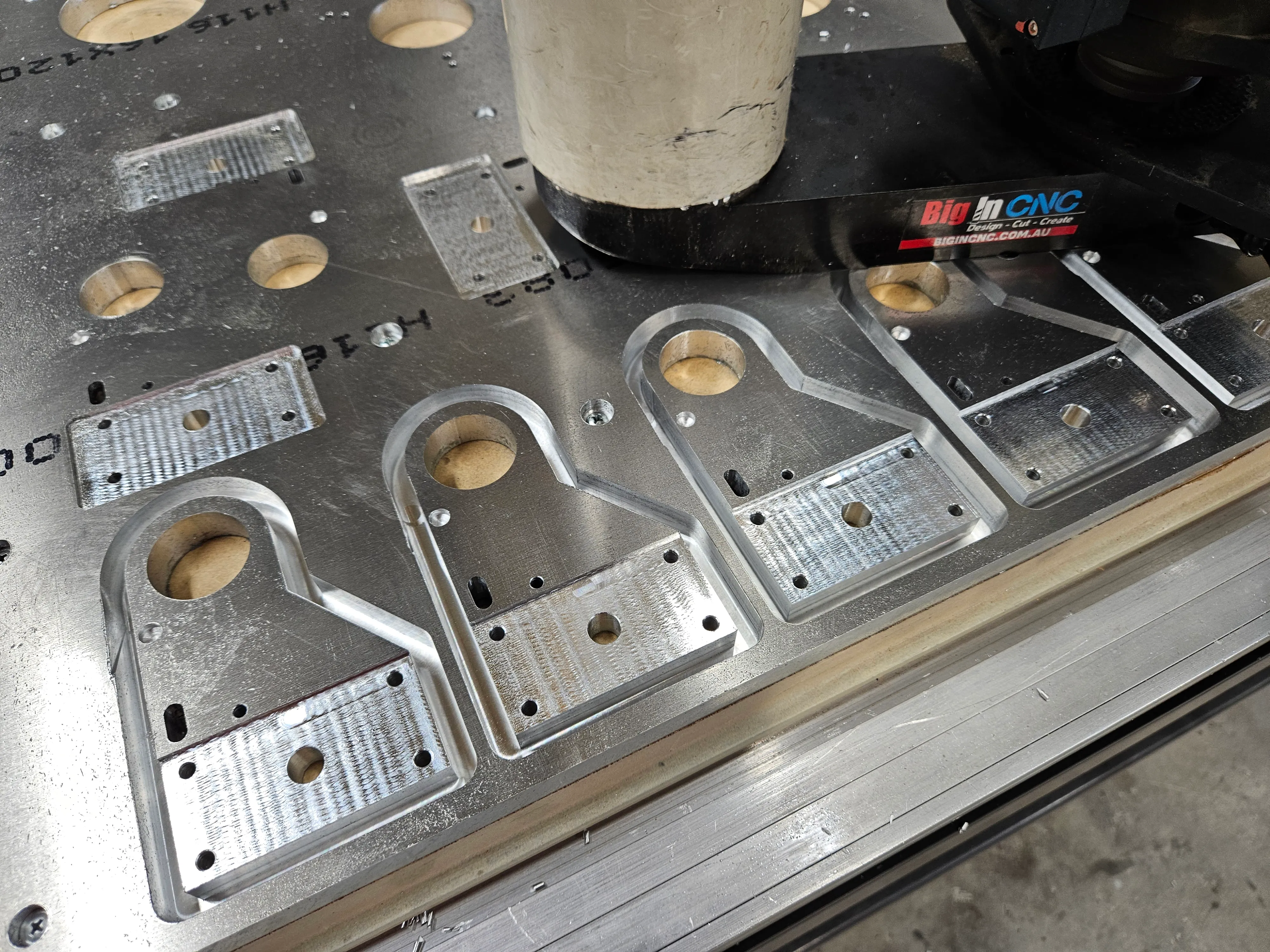 Row of five finished aluminium brackets with milled flats and mounting holes, sitting on the Multicam bed